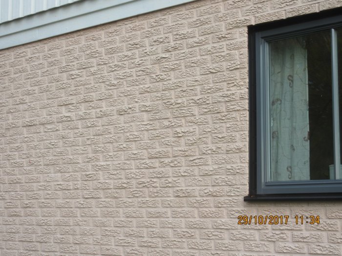 Råhuggen tegelvägg målad i rosa med tecken på vittring nära ett fönster, provyta av slätputs synlig.