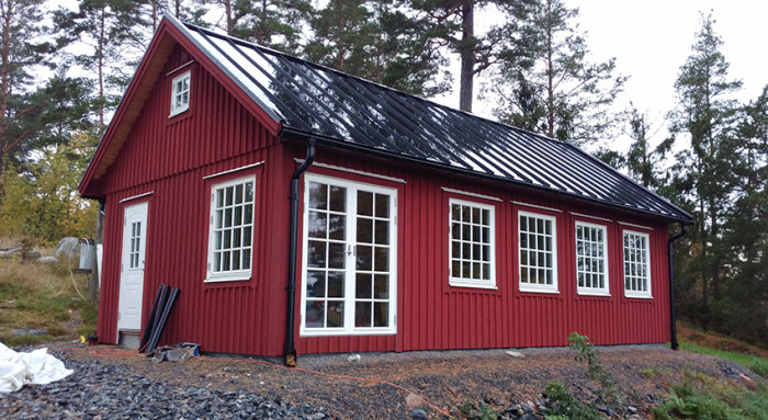 Rödfärgat hus från CA-boden med glänsande svart tak och vita fönster, omgivet av natur.