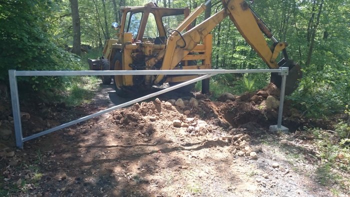 Gul grävmaskin som installerar en bom vid en grusväg i skogslandskap för att förhindra obehörig tillträde.