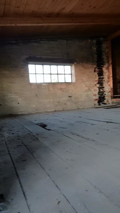 Vanha ullakko, jossa on kulunut puulattia, ikkuna ja muurattu tiiliseinä. Lattia on hieman epätasainen ja ikkunasta näkyy ulos.