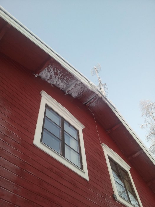 Röd husfasad med ett fönster, snötäckt tak och antenn, möjlig isskada.