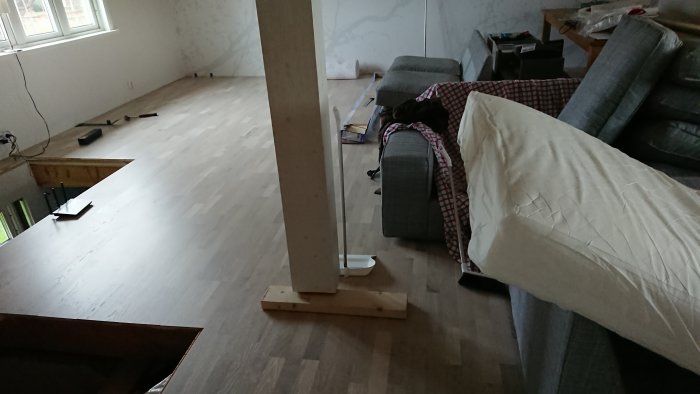 Renoverat vardagsrum med nytt ljust trägolv, oplacerade möbler och en pelare mitt i rummet.