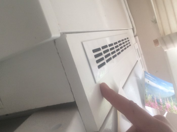 Ventilationsgaller monterat i spånskiva ovanför kylskåp med pekfingret som storleksreferens.