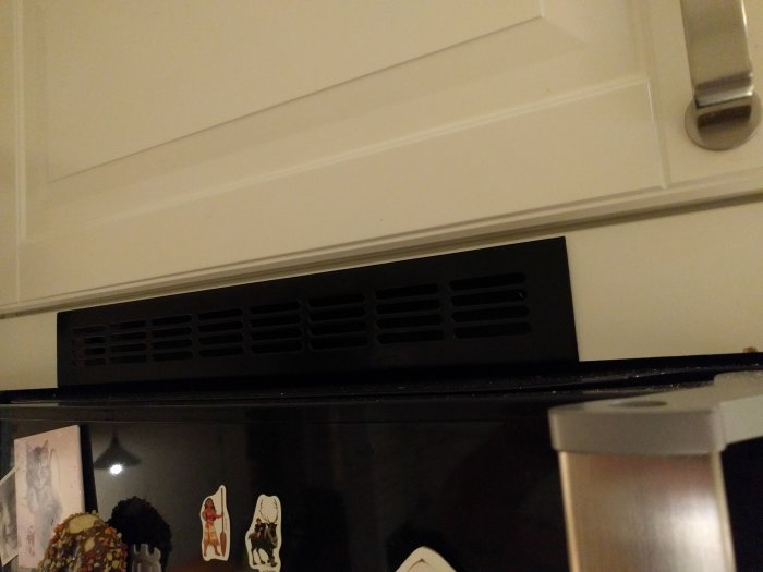 Svart ventilationsgaller ovanpå en kyl, i nivå med vit skåplucka och köksskiva.
