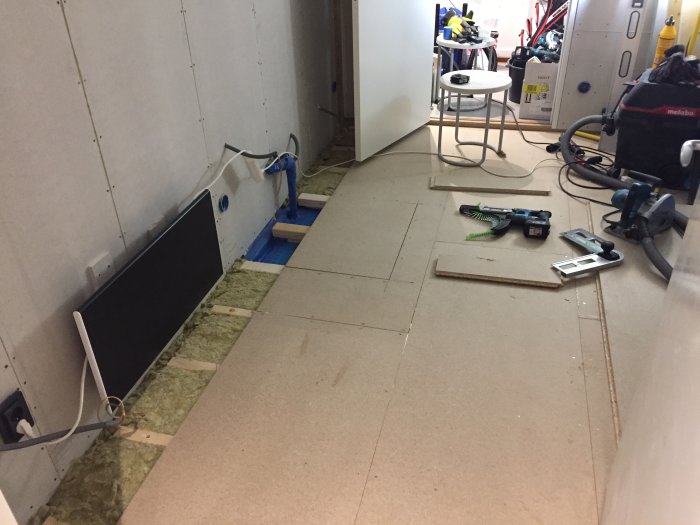 Pågående renoveringsarbete med gipsskivor på vägg och oulagt golv med verktyg och byggmaterial.
