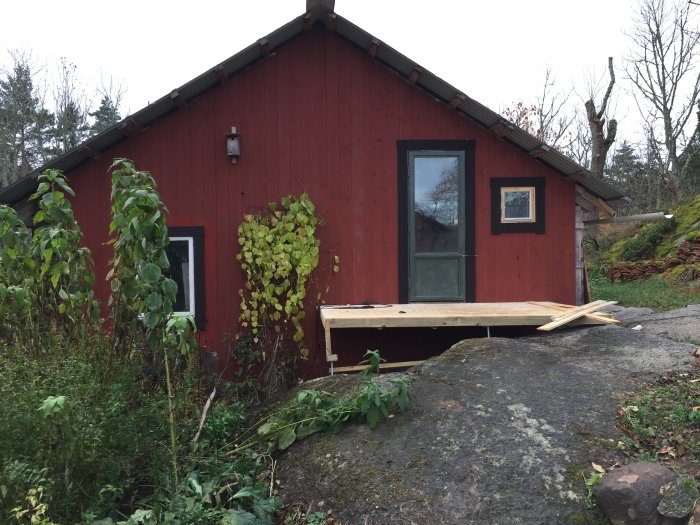 Röd stuga under renovering med nybyggd trätrall utanför, belägen på en bergknalle omgiven av grönska.