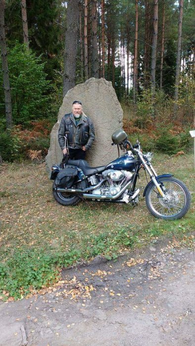 Man i läderjacka lutar sig mot en motorcykel framför en sten i skogen.