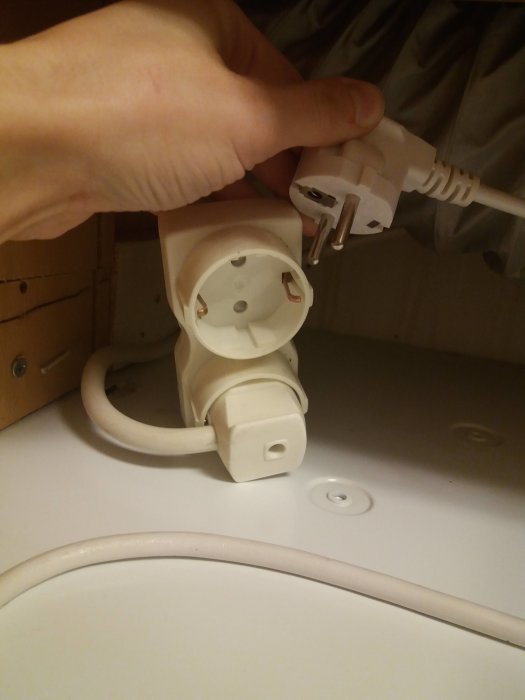 Hand som håller en elsladd bredvid en opolerad, utanpåliggande dubbel eluttag på en vit yta.