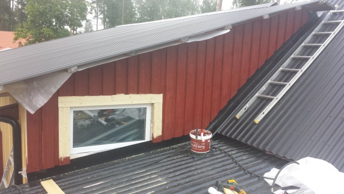 Del av ett rött garage under renovering med öppet fönster och en steg på det svarta taket, bredvid en burk rödfärg.