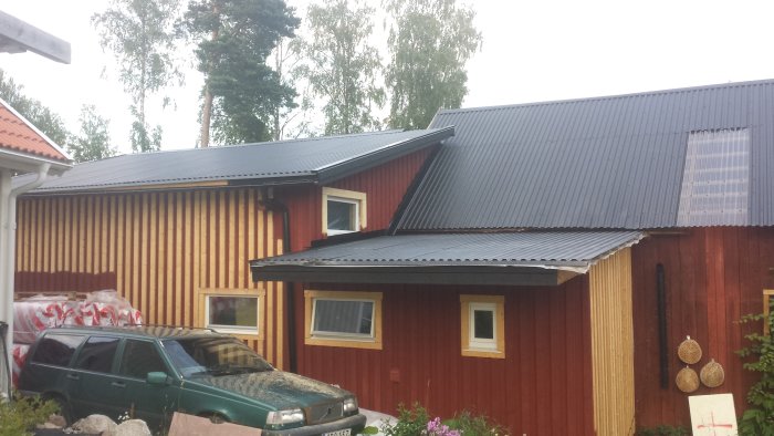 Trägarage med röd och naturfärgad panel och svart wellplåttak, bredvid en grön bil och byggmaterial.