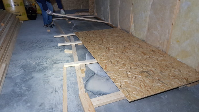 Improvised wooden bridge over unevenly cured self-leveling compound on garage floor with visible water and construction materials.