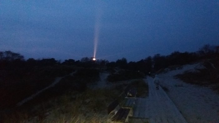 Trästig i sanddynlandskap vid skymning med tänd fyr i bakgrunden som lyser upp en stråle i luften.