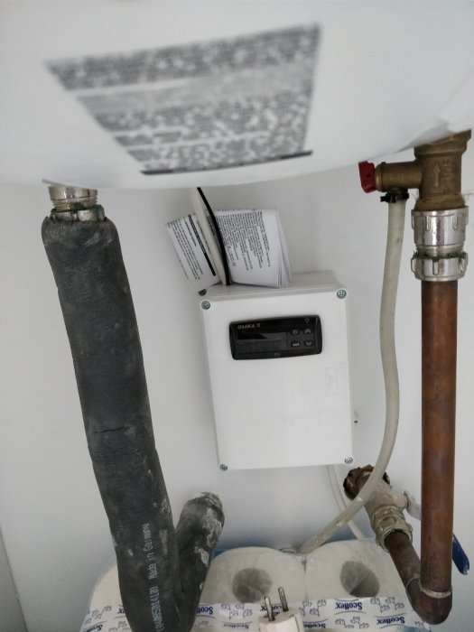 En värmeregulator installerad på en vit vägg med synliga rörkopplingar och en instruktionsmanual öppen bakom den.