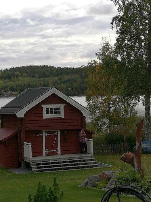 Rödmålat härbre med vitknutade hörn står nära vatten, person lutar sig mot veranda, omgivet av grönska.