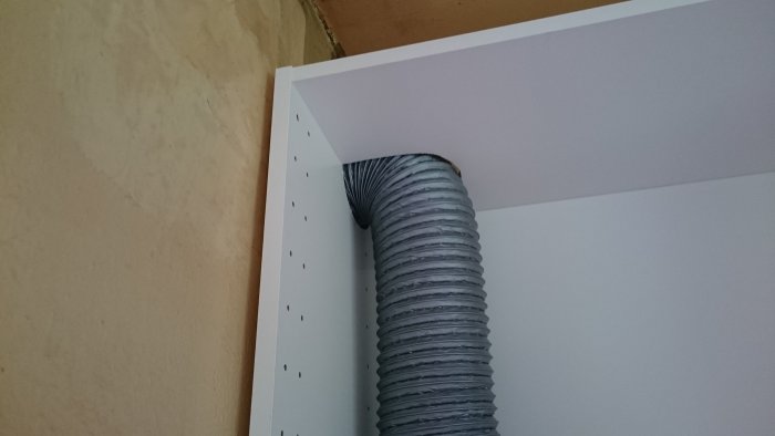 Flexibel ventilationsslang som går genom öppningen i ett vitt IKEA köksöverskåp under installation.