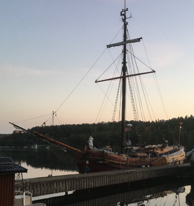 Segelbåt förtöjd vid kaj i skymningen, stilla vatten och skog i bakgrunden.