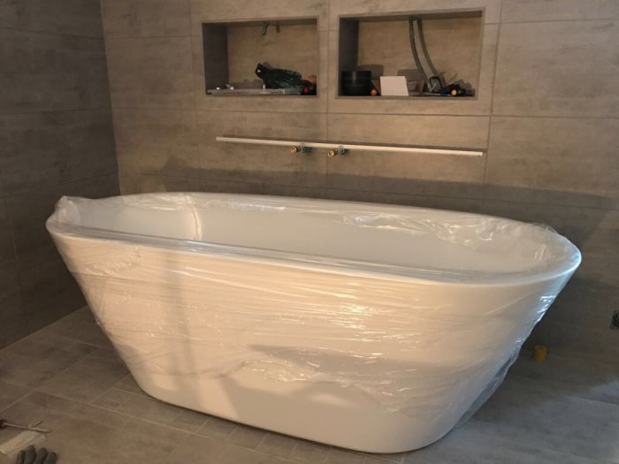 Nytt badkar inplastat under installation i badrum med kakelväggar och tvättställ i bakgrunden.