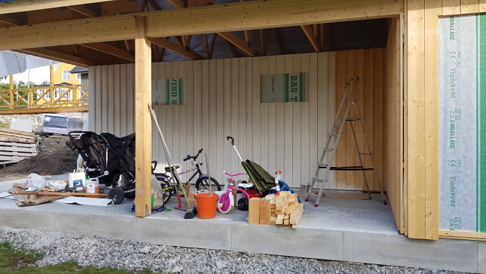 Halvfärdigt carport med synliga reglar och vindskydd, liggande läkter, barnleksaker och en stege framför.