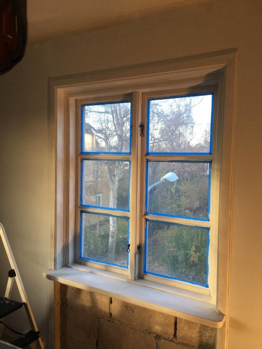 Nymålade vitgrundade fönsterkarmar med blå tejp runt glaset i en ljus rumsmiljö.