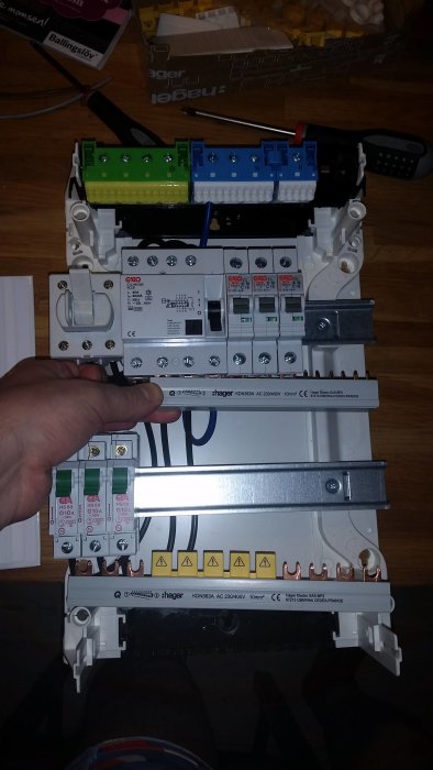 Hand håller två elektriska fasskenor intill en öppen strömbrytarcentral med märkningar.