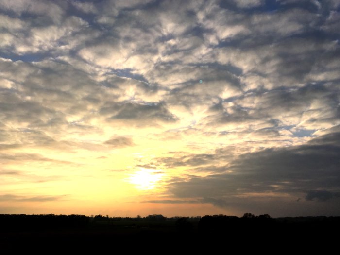 Dramatisk himmel vid solnedgång med mönstrade moln och varma färger över ett silhuetterat landskap.