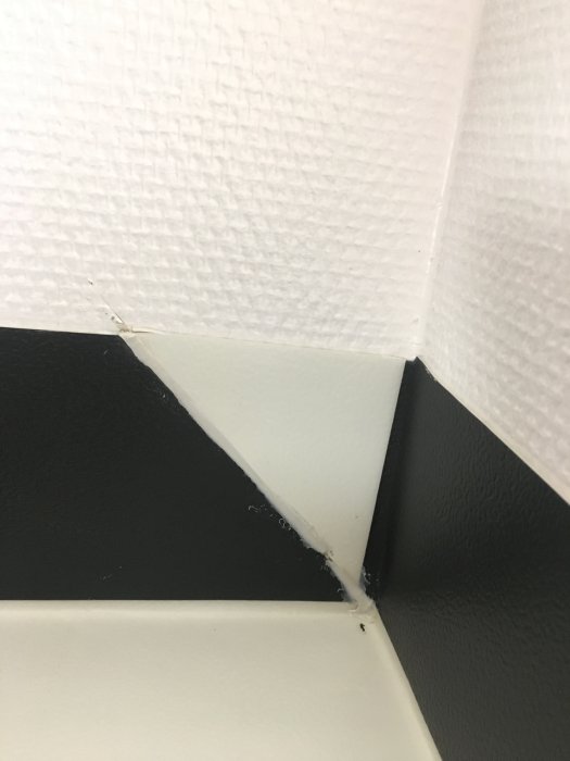 Hörnet av ett badrum med dåligt utförda svetsningar och missfärgningar på en ny svart golvmatta.