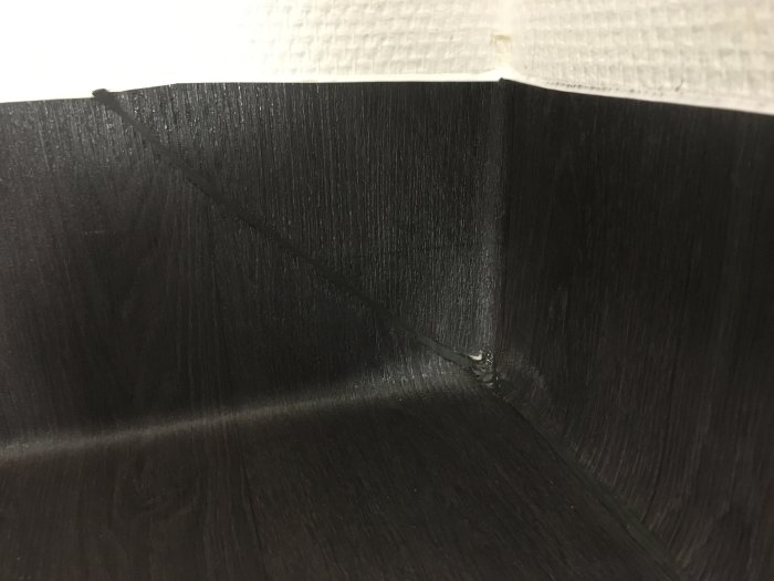 Svart golvmatta med synliga svetsfogar och ojämnheter vid hörnet och vägganslutningen.