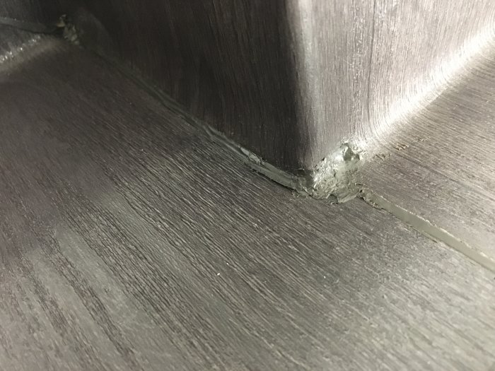 Svart golvbeläggning vid hörn med dåligt utförda svarta svetsningar och ojämnheter.