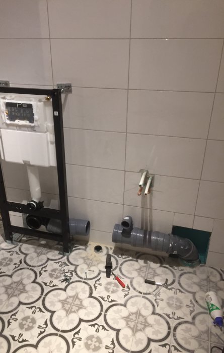 Renovering av badrum med synlig vägghängd toalettstomme och rörinstallationer på kaklad vägg och mönstrat golv.