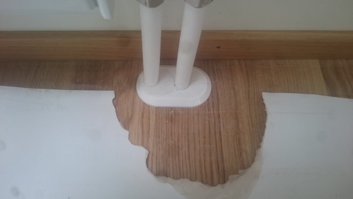 Vit täckbricka runt två rör på trägolv med ojämna kanter där vägg möter golv.