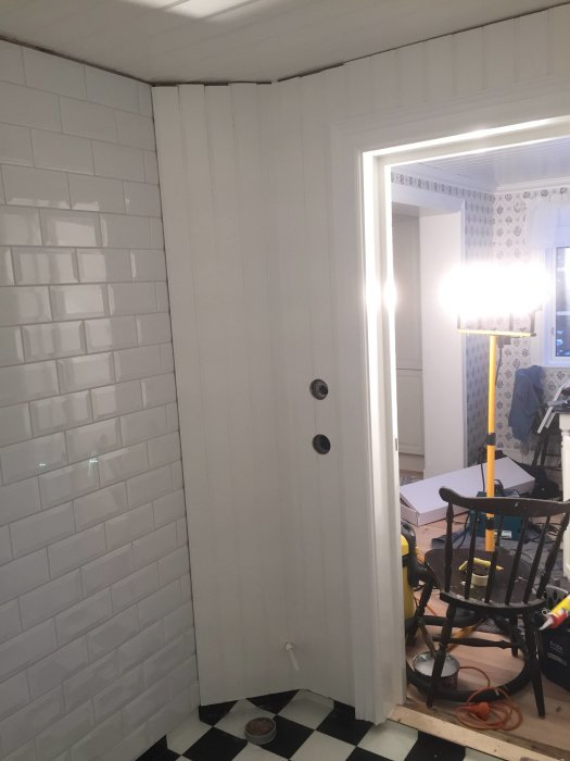 Ett badrumshörn med vita kakelväggar och svartvit rutig golvmatta under renovering, verktyg synliga.