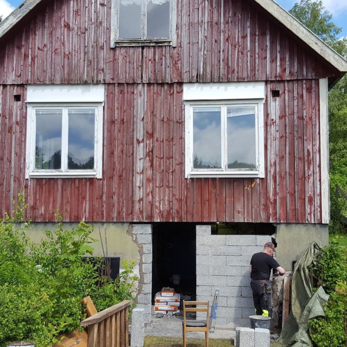 Renovering av fasad på äldre hus, person målar vägg, slitna brädor och öppen källare syns.