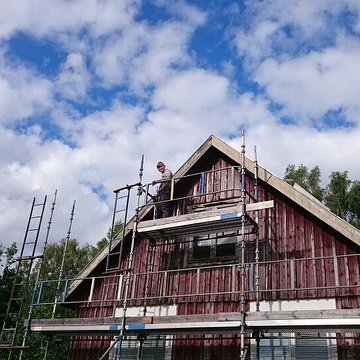 Renovering av husfasad med person på byggnadsställning och rödmålade träväggar under arbete.