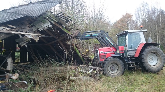 Röd traktor med frontlyft river ett förfallet stall, skräp och träplankor syns.