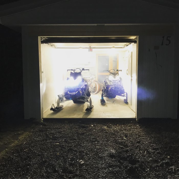 Två snöskotrar parkerade i ett upplyst garage, med öppen port och en cykel i bakgrunden.