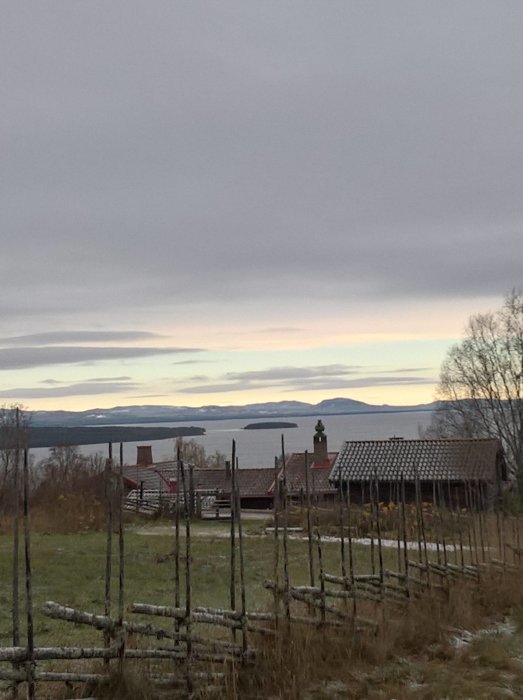 Molnig himmel över landskap med sjö, Gesunda i bakgrunden, och en midsommarstång på en tom snöfri mark.