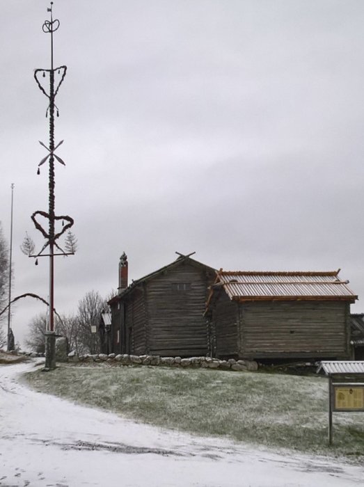 Midsommarstång med snötäcke framför en timrad stuga, snö på marken, disig himmel.