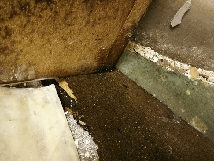 Hörn av ett källargolv med synlig betong, delar av gamla golvmaterial och nedbrutet material, behov av renovering.