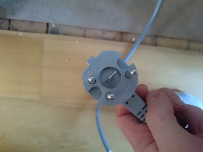 Hand håller en grå elektrisk spiskontakt mot ett trägolv.