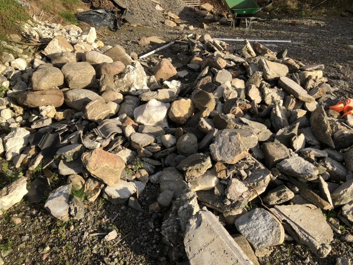 Hög med blandade stenar och byggavfall på marken med gröna skott och grushög i bakgrunden.