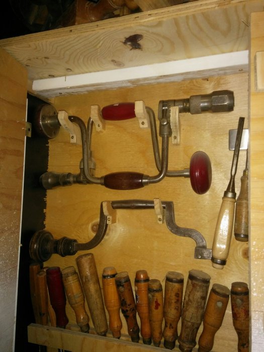 Samling av nio gamla handborrar med trähandtag upphängda på en vägg, inklusive en elektrikerborr.