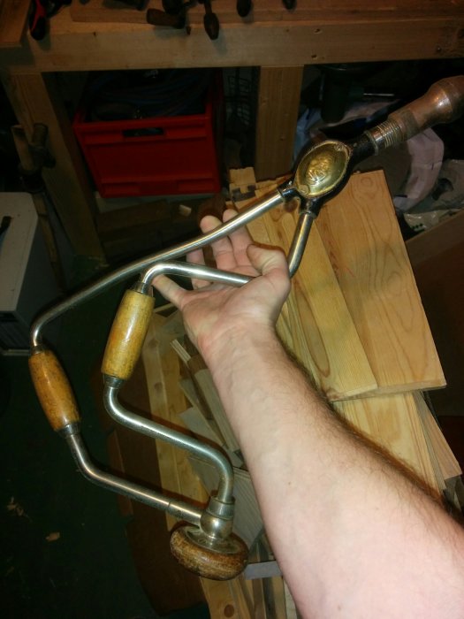 Hand håller en gammal manuell borr med trähandtag framför en verkstadsbänk.