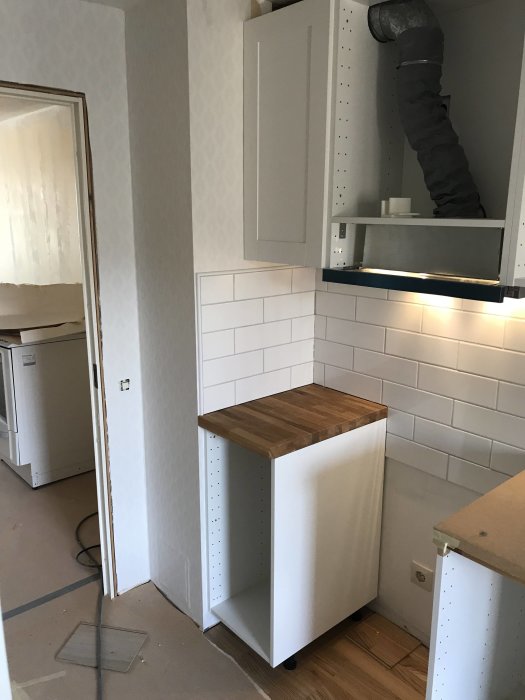 Kök under renovering med vit kakelvägg, skåp och träarbetsyta i dagsljus.