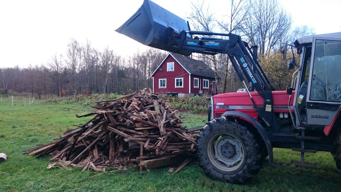 Traktor med frontlastare som tömmer en hög avvecklat trävirke på en gräsmatta framför ett rött hus.