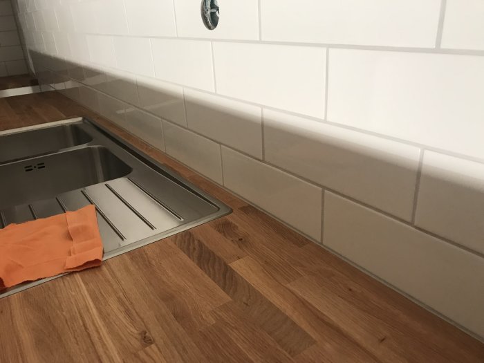 Hörn av nyinstallerat kök med träbänkskiva, rostfri diskho och vitt kakel, detalj av mjukfog längs sockeln.