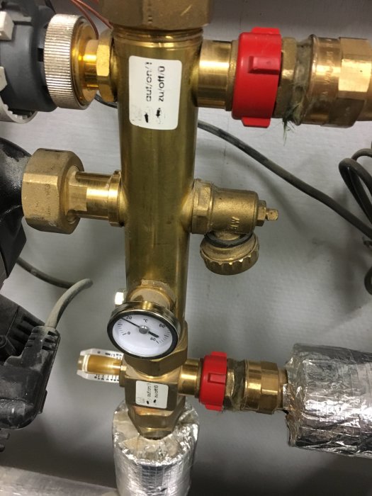 Mässingsfärgad värmesystemkomponent med justerbar röd ring och anslutna mätare, belägen i en teknisk installation.