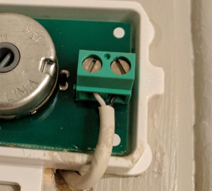 Två kablar anslutna till ett grönt kopplingsblock på en termostat med omgivande vit vägg.