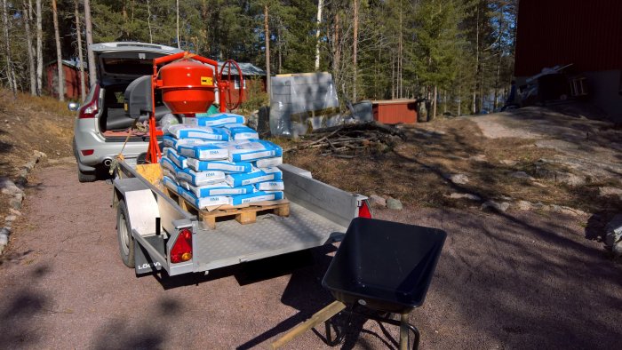 Släpvagn fullastad med säckar av grovbetong parkerad nära skog och röd byggnad.