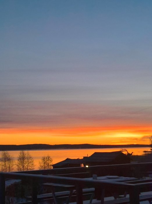 Soluppgång över en sjö med silhuetter av hus och träd mot en orangehimmel.