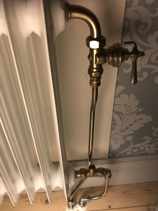 Ny lenhovdaradiator kopplad med mässingsventiler och rör mot en vit vägg med blommigt tapetmönster.